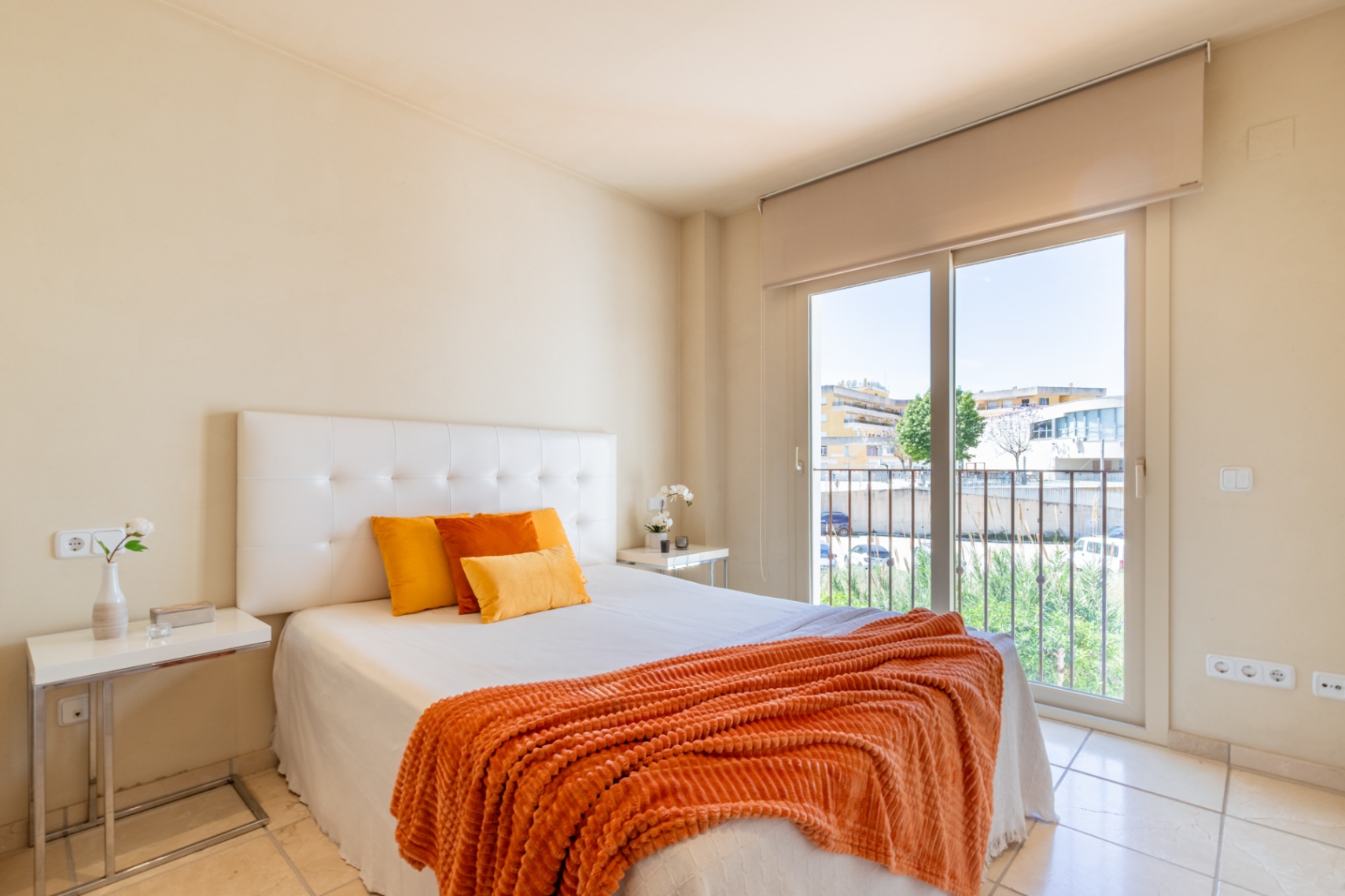 Chambre lumineuse avec lit double, tête de lit capitonnée, sol carrelé clair et accès direct à un balcon offrant des vues urbaines.