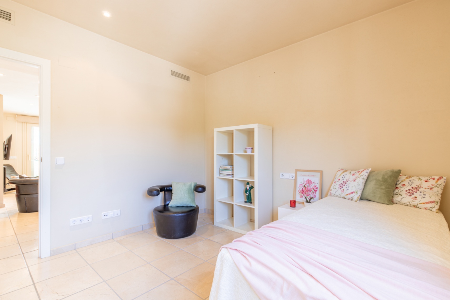 Chambre lumineuse avec un lit double, carrelage clair et murs crème. Équipée d'une étagère blanche, d'un fauteuil foncé et d'une ouverture vers une pièce contiguë.