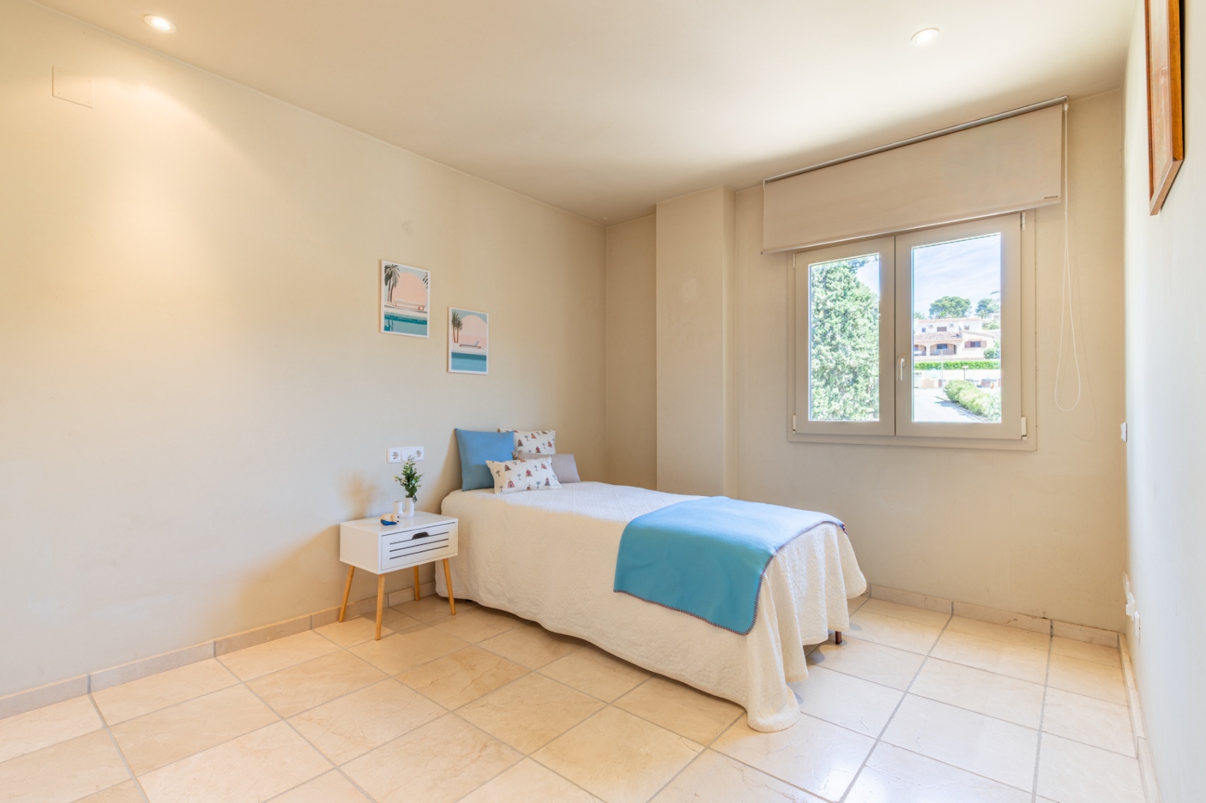 Chambre secondaire lumineuse avec un lit simple aux draps bleus et blancs, carrelage clair et fenêtre offrant une vue sur la verdure et les résidences. Éclairage encastré au plafond.