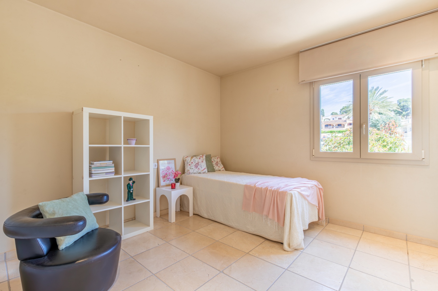 Chambre secondaire lumineuse avec un lit simple, une étagère blanche et un fauteuil sombre. La fenêtre offre une vue sur la verdure et une lumière naturelle abondante sur le sol carrelé.