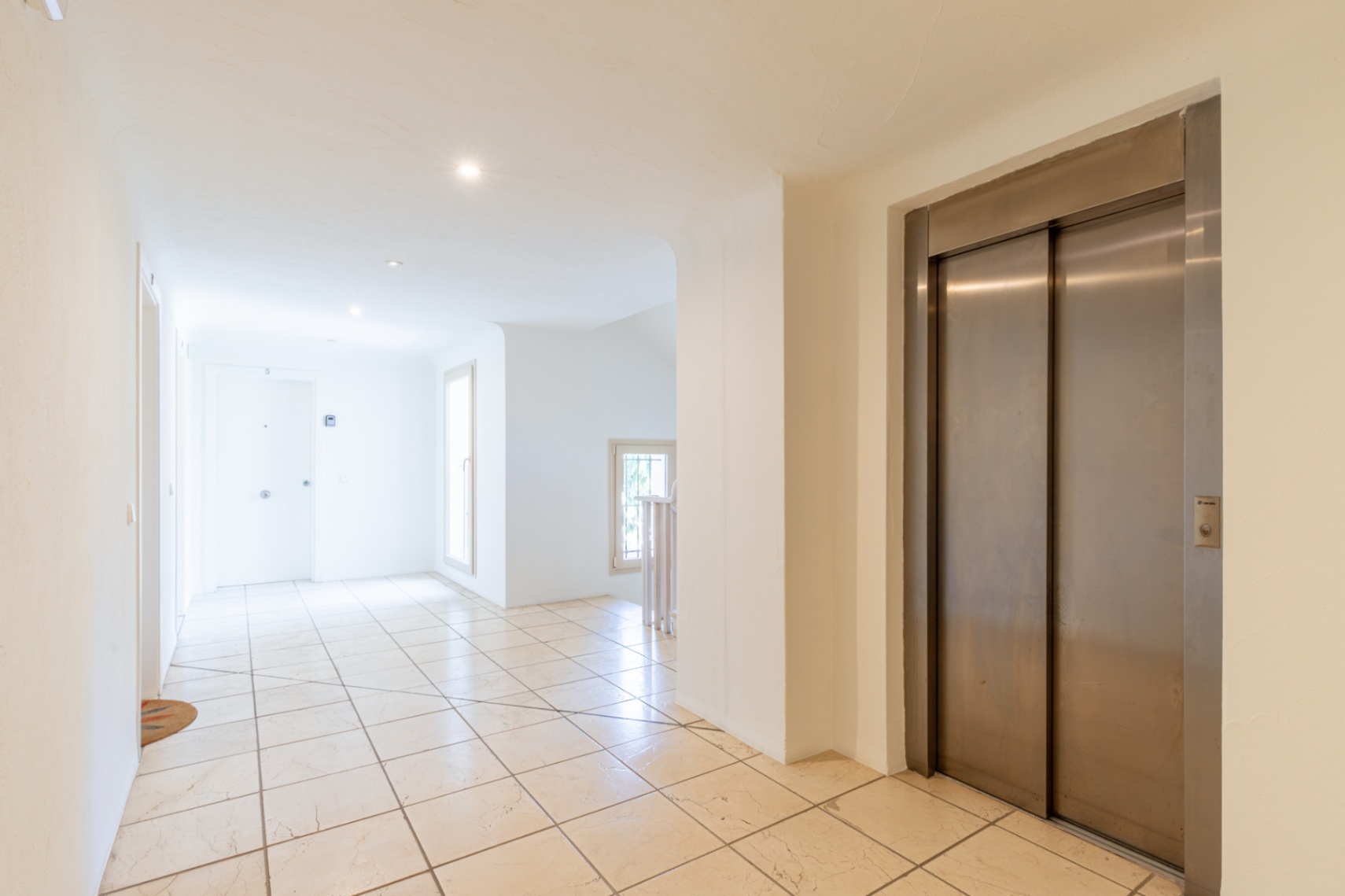 Couloir commun lumineux avec carrelage clair, ascenseur en acier inoxydable et lumière naturelle des fenêtres. Éclairage encastré au plafond.