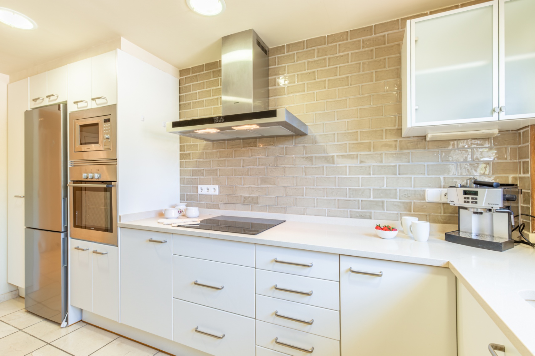 Cuisine contemporaine avec des armoires blanches épurées, des appareils encastrés en acier inoxydable et un dosseret distinctif en carrelage beige effet brique.