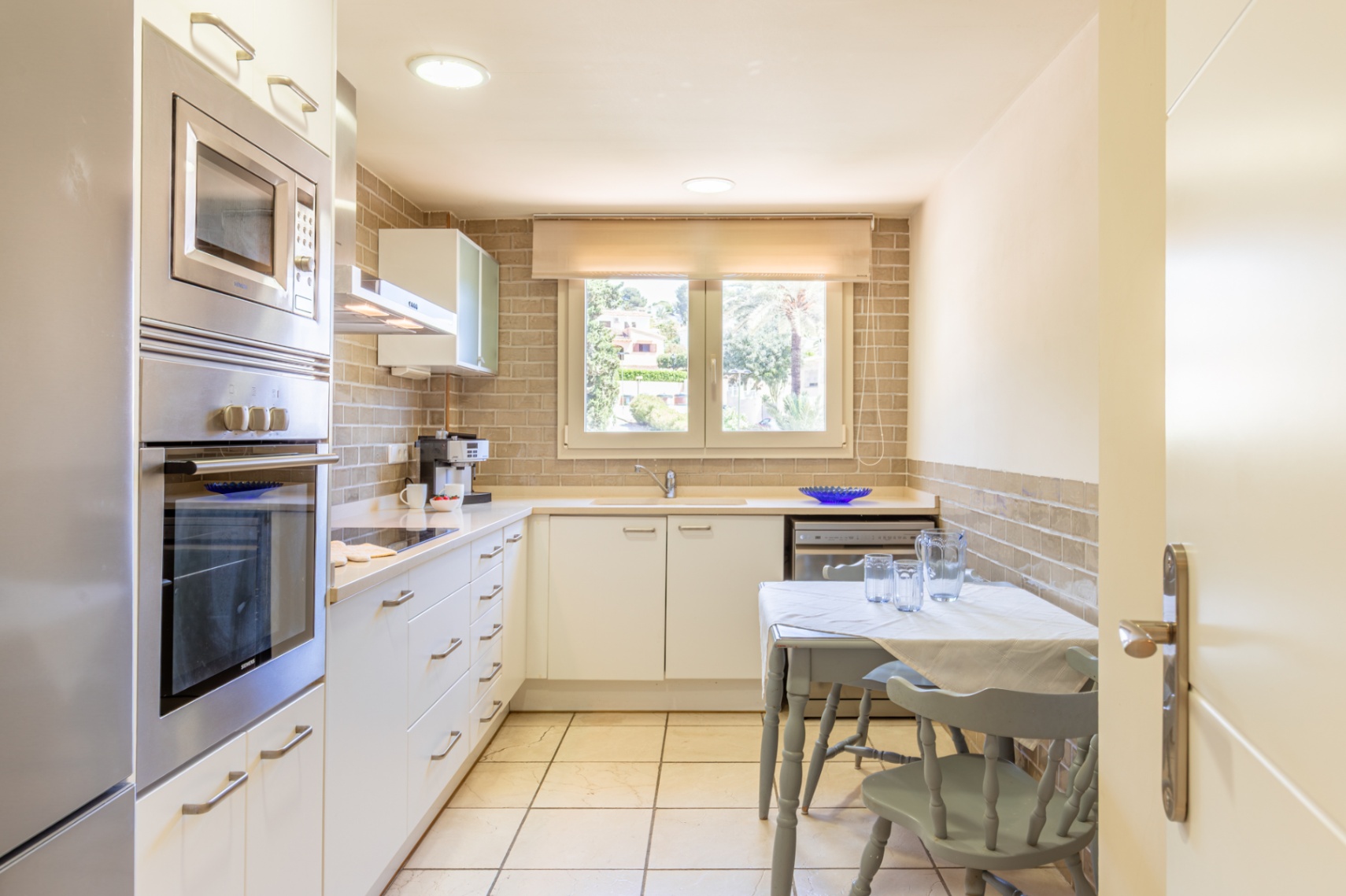 Cuisine fonctionnelle avec électroménager intégré en acier inoxydable, plan de travail clair et crédence en briques. Comprend une table compacte et une fenêtre sur verdure.