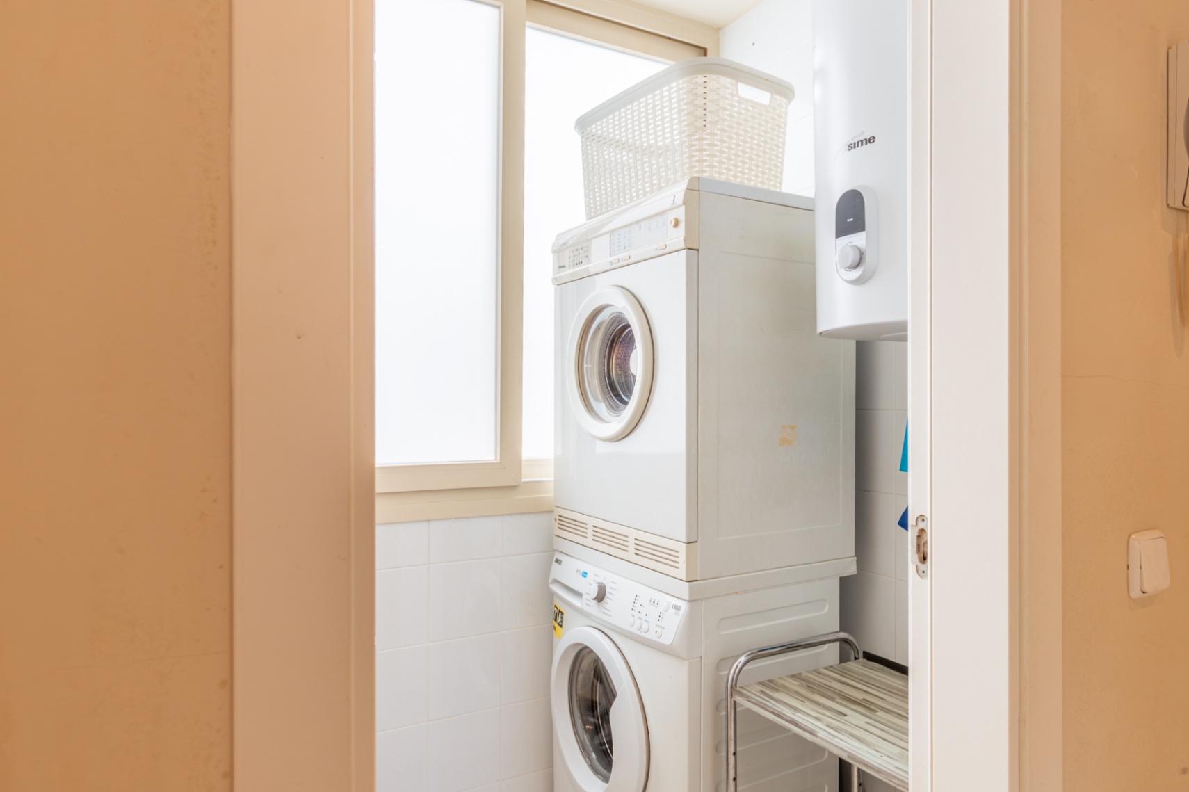 Espace buanderie compact avec lave-linge et sèche-linge superposés, optimisant l'espace. Chauffe-eau intégré et fenêtre dépolie pour une lumière naturelle.