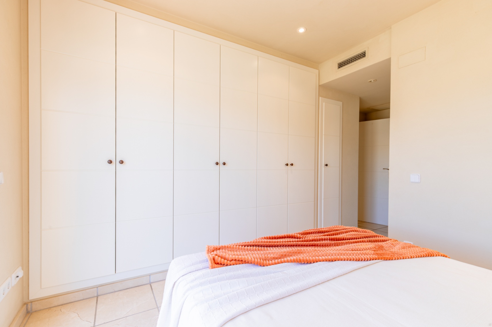 Grande armoire encastrée aux portes blanches à panneaux et poignées sombres, offrant un rangement généreux dans cette chambre lumineuse avec carrelage au sol.