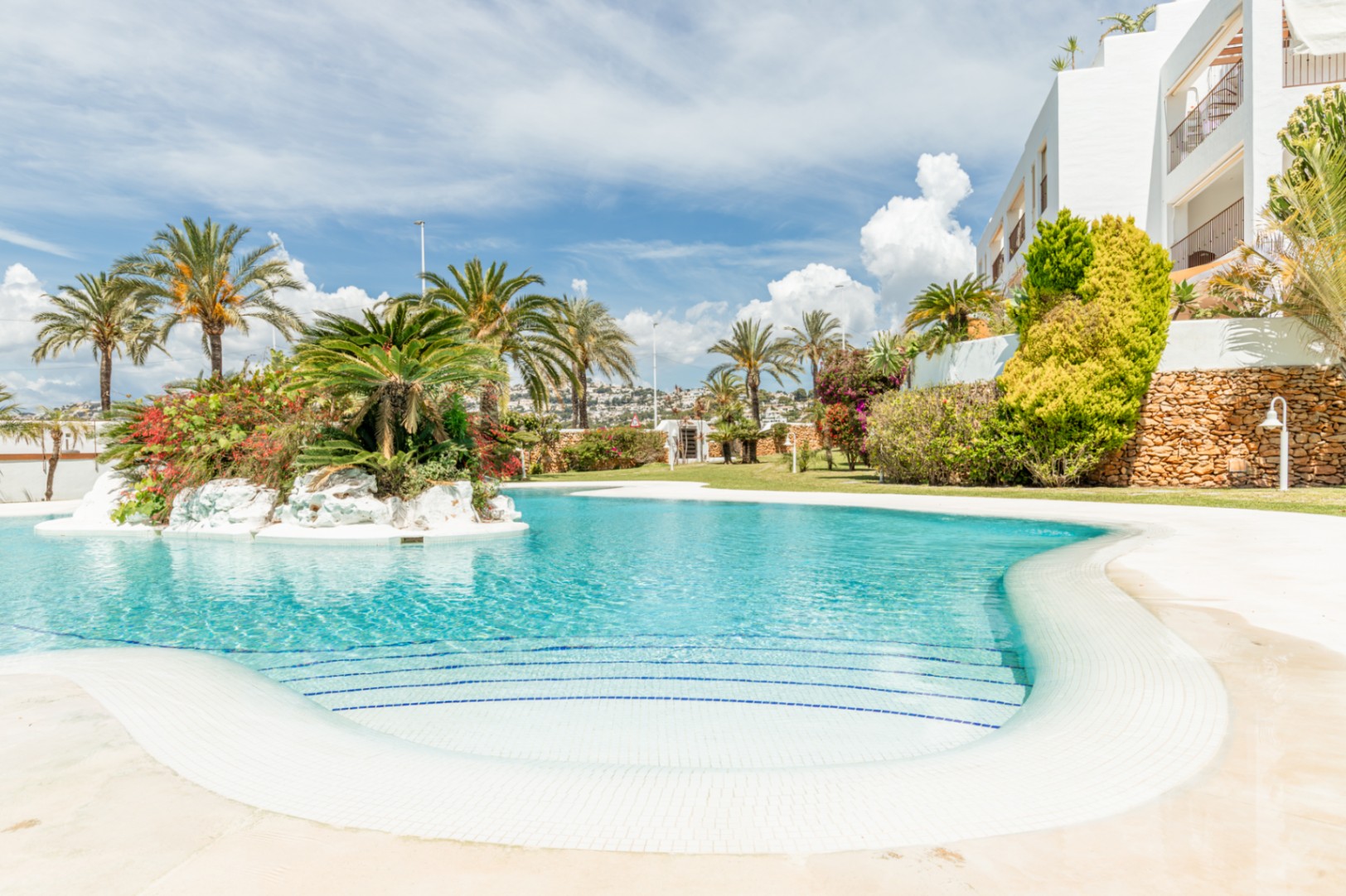 Grande piscine communautaire aux courbes élégantes, avec escaliers en mosaïque blanche et bleue, et une île centrale luxuriante de palmiers et fleurs.