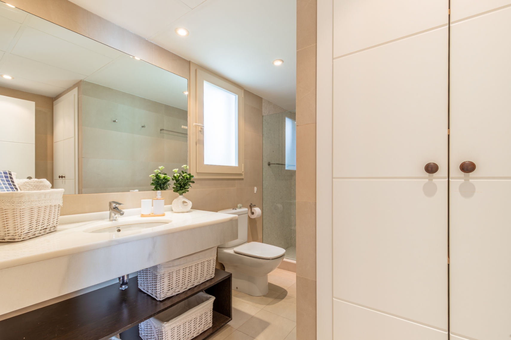 Salle de bain complète avec lavabo en marbre, grand miroir et rangements intégrés. Douche avec paroi vitrée, fenêtre discrète et touches décoratives. Tons neutres, grande capacité de rangement.