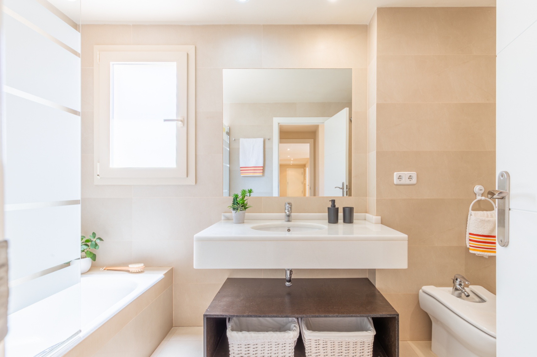 Salle de bain complète avec meuble-lavabo suspendu, vasque intégrée et grand miroir. Carrelage beige clair, fenêtre discrète et baignoire encastrée.