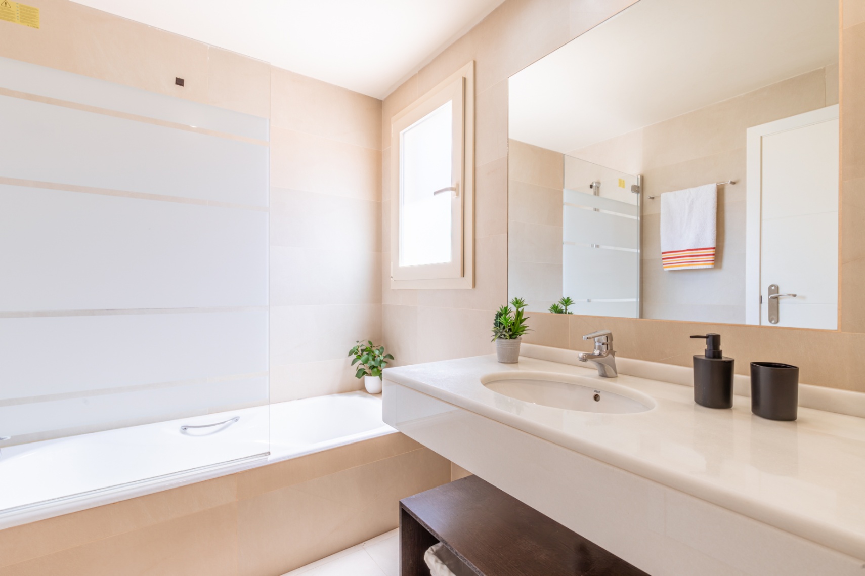 Salle de bain moderne avec meuble-lavabo flottant blanc, grand miroir et baignoire avec pare-douche en verre rayé, sur des murs carrelés neutres.