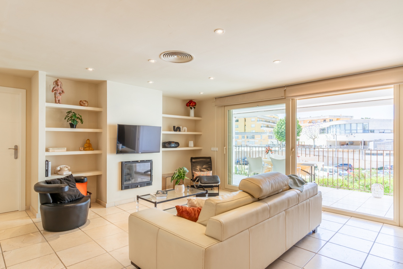 Salon lumineux avec canapé crème, carrelage et étagères intégrées autour d'une cheminée et TV. De larges baies vitrées s'ouvrent sur une terrasse privée avec vue urbaine.