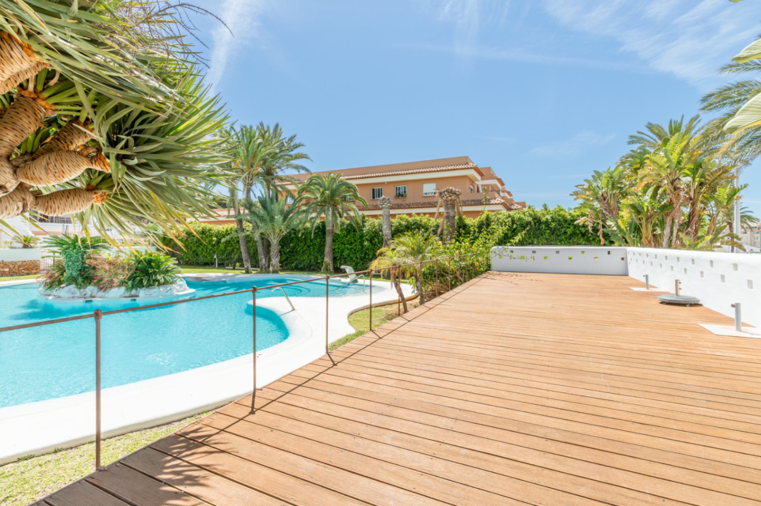 Vaste espace commun avec une grande piscine incurvée et un îlot paysager central. La terrasse en bois adjacente offre un large espace, entourée de palmiers et de végétation méditerranéenne luxuriante.