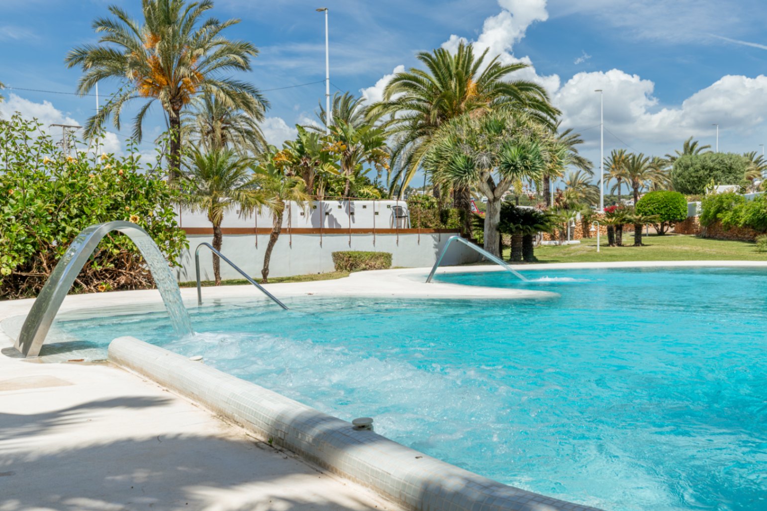 Vaste piscine communautaire avec jets d'eau et zones bouillonnantes, entourée de palmiers luxuriants et de verdure sous un ciel bleu éclatant.