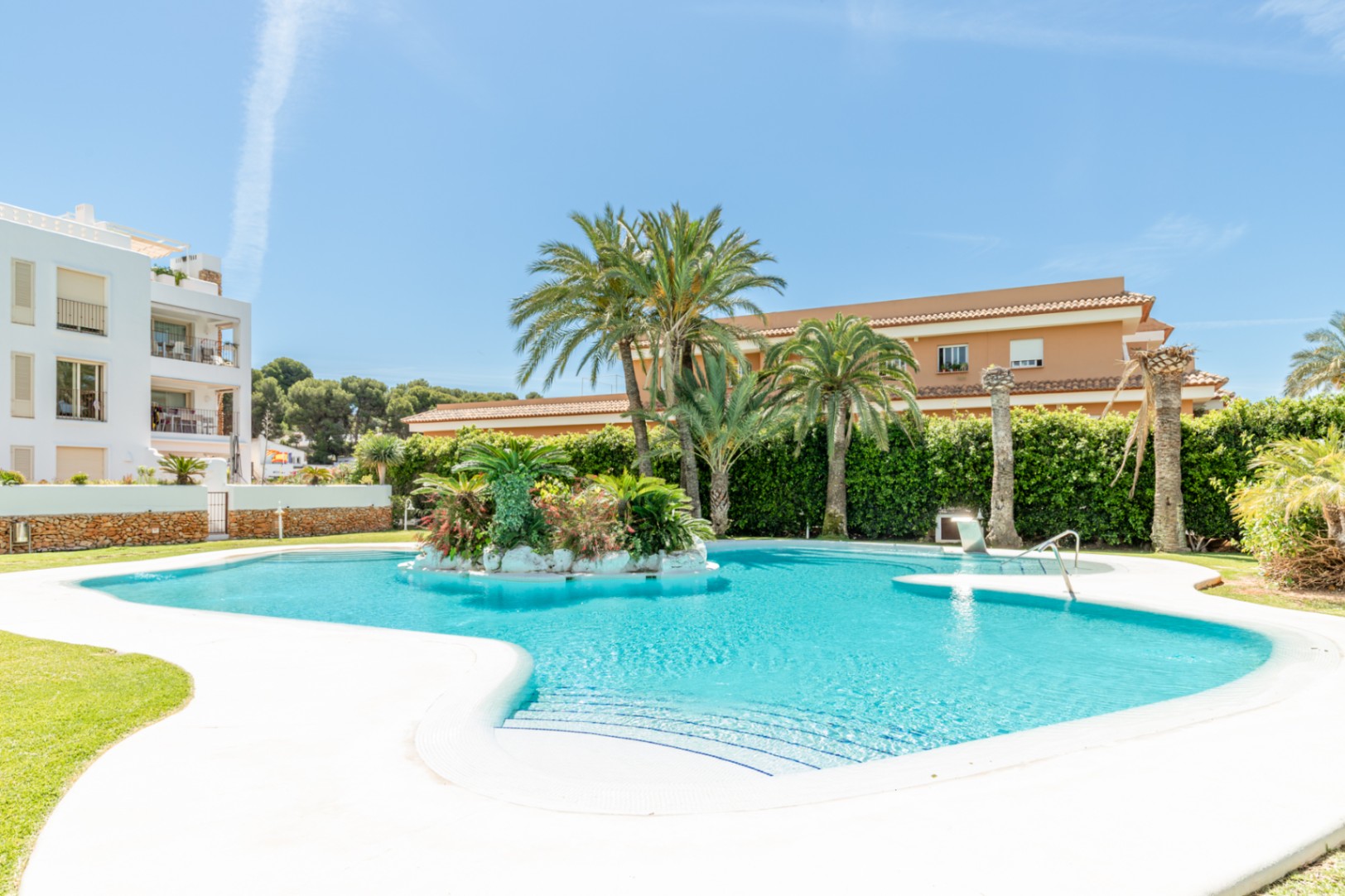 Vaste piscine communautaire de forme irrégulière avec une île centrale luxuriante de palmiers et végétation, entourée de verdure et de bâtiments sous un ciel clair.