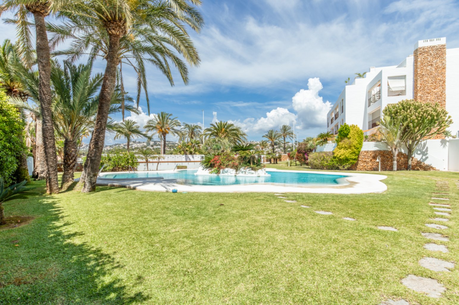 Vaste piscine commune avec un îlot végétalisé intégré, entourée de pelouses impeccables et de palmiers matures. Le bâtiment résidentiel blanc aux accents de pierre est visible sous un ciel clair.