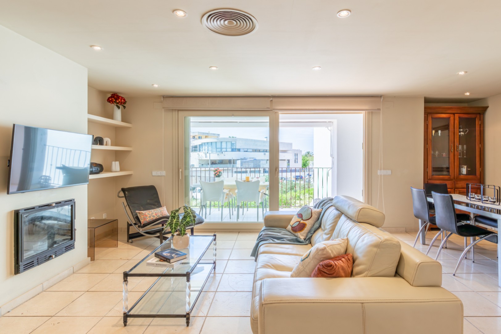 Vaste salon avec canapé en cuir crème, cheminée intégrée et TV murale. De grandes portes coulissantes mènent à une terrasse privée, offrant lumière naturelle et fluidité.