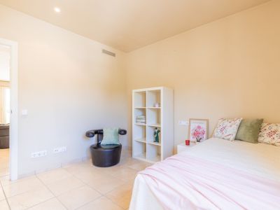 Chambre lumineuse avec un lit double, carrelage clair et murs crème. Équipée d'une étagère blanche, d'un fauteuil foncé et d'une ouverture vers une pièce contiguë.