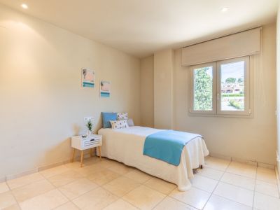Chambre secondaire lumineuse avec un lit simple aux draps bleus et blancs, carrelage clair et fenêtre offrant une vue sur la verdure et les résidences. Éclairage encastré au plafond.