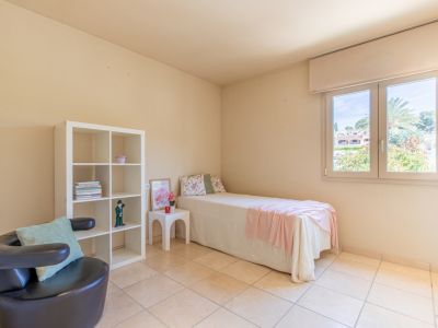 Chambre secondaire lumineuse avec un lit simple, une étagère blanche et un fauteuil sombre. La fenêtre offre une vue sur la verdure et une lumière naturelle abondante sur le sol carrelé.