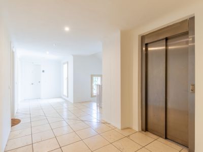 Couloir commun lumineux avec carrelage clair, ascenseur en acier inoxydable et lumière naturelle des fenêtres. Éclairage encastré au plafond.