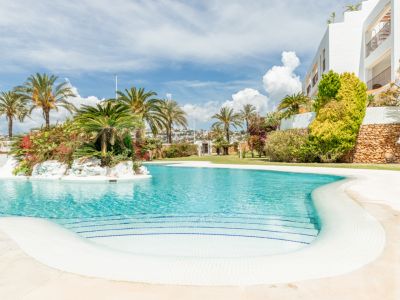Grande piscine communautaire aux courbes élégantes, avec escaliers en mosaïque blanche et bleue, et une île centrale luxuriante de palmiers et fleurs.