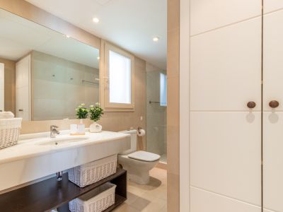 Salle de bain complète avec lavabo en marbre, grand miroir et rangements intégrés. Douche avec paroi vitrée, fenêtre discrète et touches décoratives. Tons neutres, grande capacité de rangement.