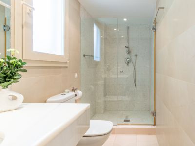 Salle de bain contemporaine avec WC, lavabo à plan blanc et fenêtre. La douche vitrée est revêtue de mosaïque et de carrelage beige, baignée de lumière naturelle.