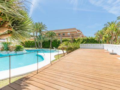Vaste espace commun avec une grande piscine incurvée et un îlot paysager central. La terrasse en bois adjacente offre un large espace, entourée de palmiers et de végétation méditerranéenne luxuriante.