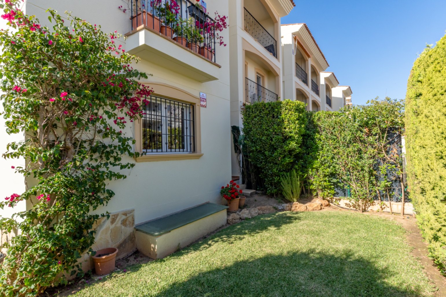 Aperçu du jardin privé avec pelouse verte, bougainvillier fleuri sur la façade et haies. Fenêtre sécurisée et banc extérieur visible.