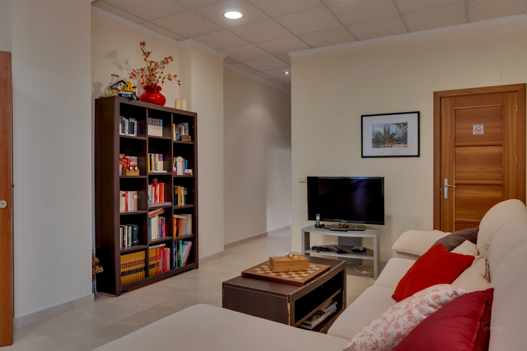 Ce salon secondaire polyvalent comprend un canapé confortable, une table basse avec échiquier et une grande bibliothèque garnie de livres. Une télévision et un sol carrelé clair sont visibles.