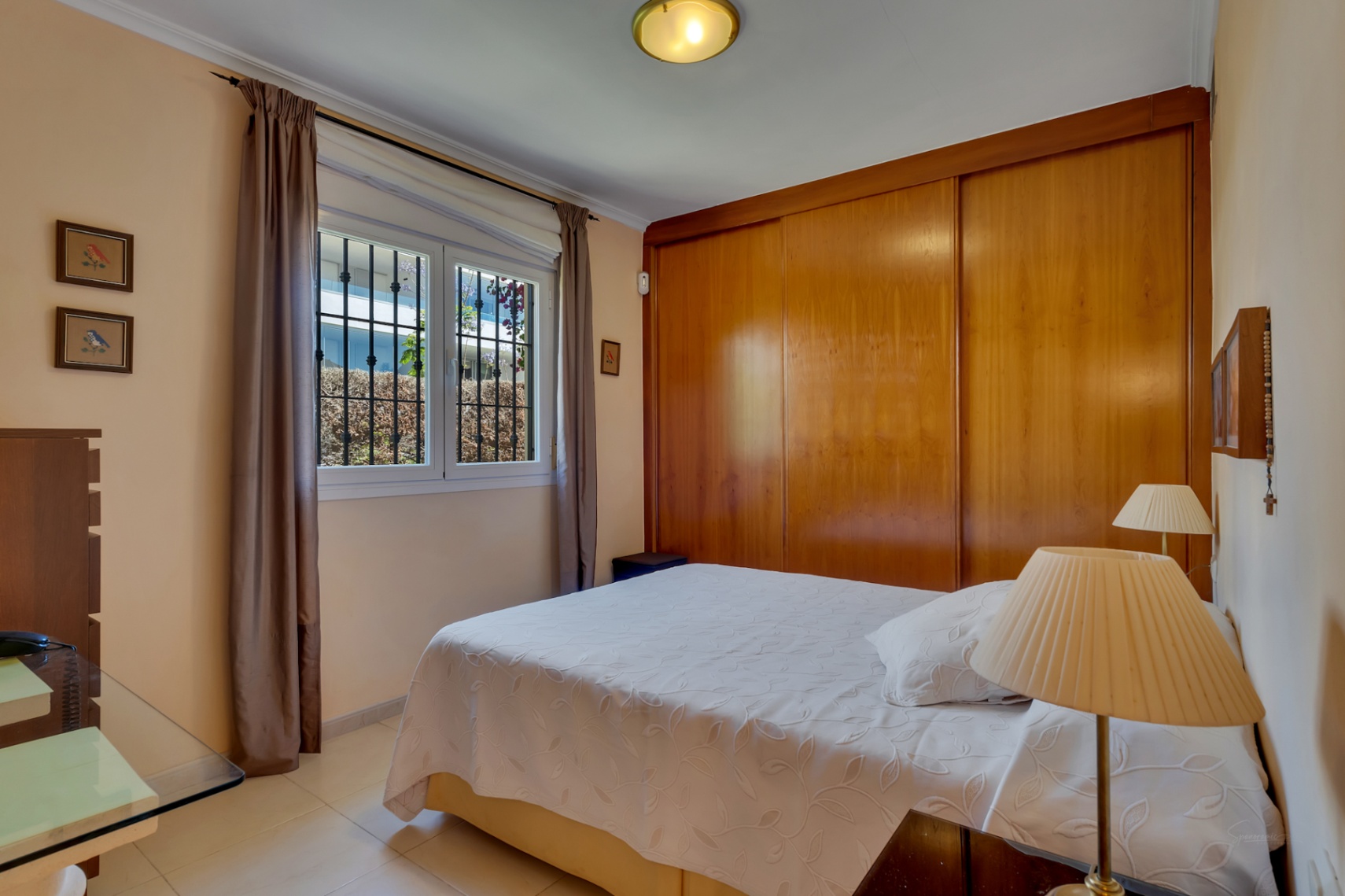 Chambre avec lit double, armoire intégrée en bois et fenêtre grillagée donnant sur la verdure extérieure.
