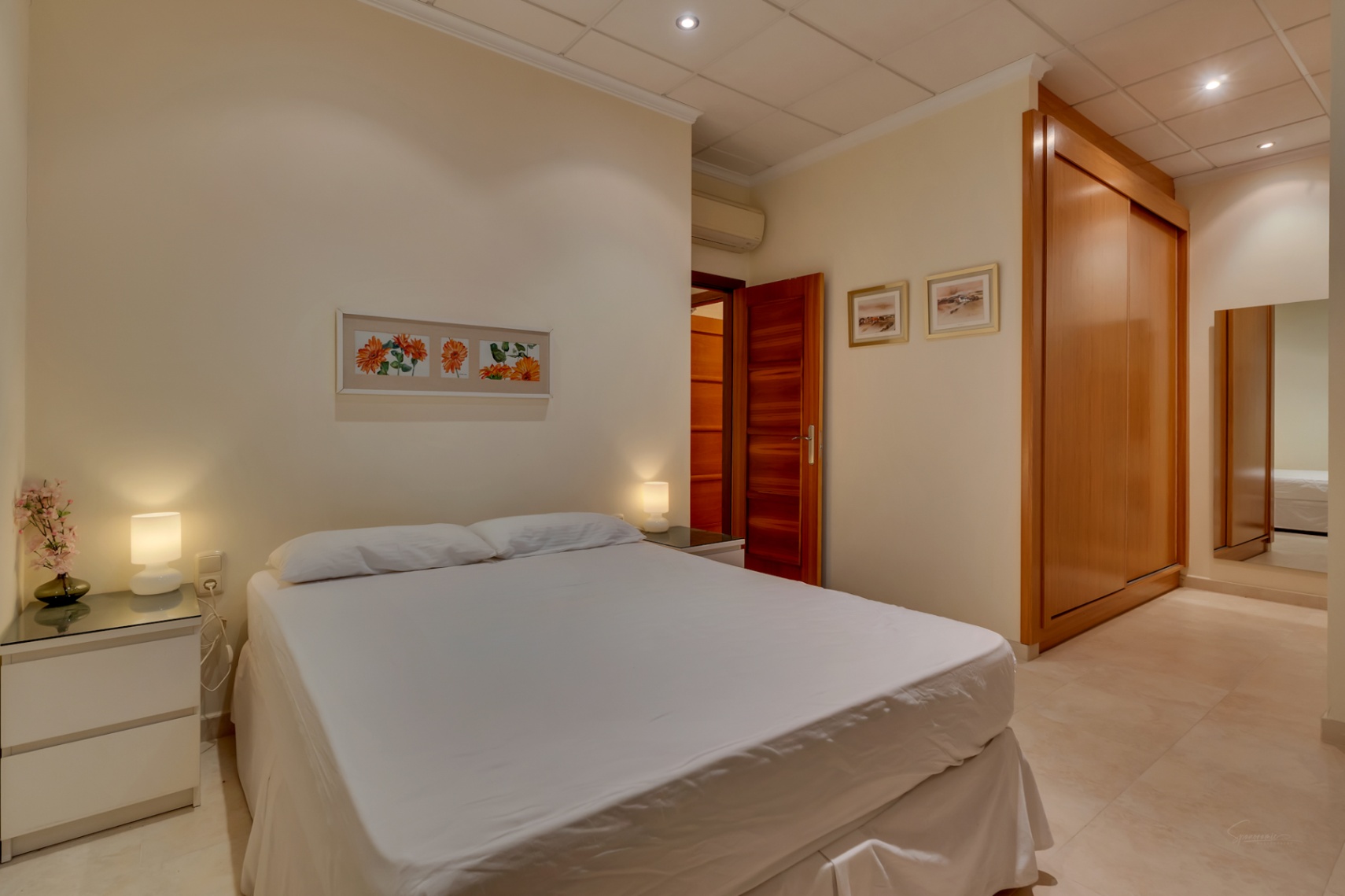 Chambre confortable avec lit double, tables de chevet modernes, grande armoire encastrée en bois et carrelage clair, équipée de la climatisation.