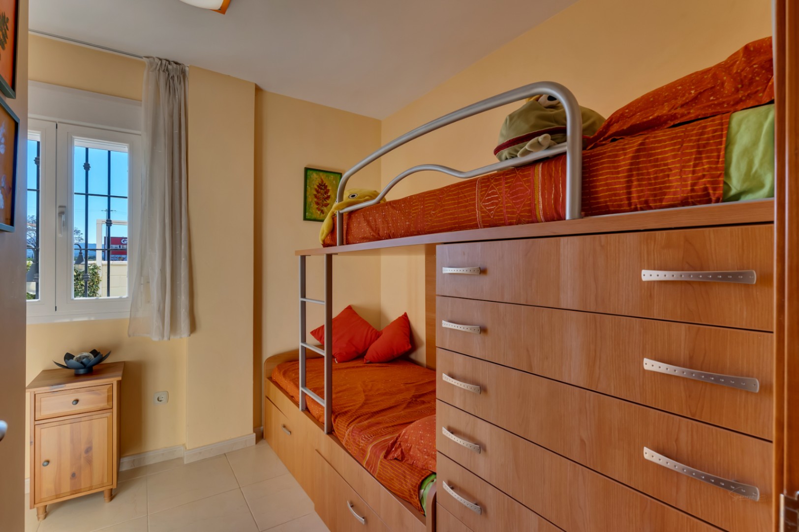 Chambre d'enfants avec lits superposés intégrés dans un meuble en bois clair, incluant des tiroirs de rangement. Une fenêtre avec grille et voilage apporte une lumière naturelle.
