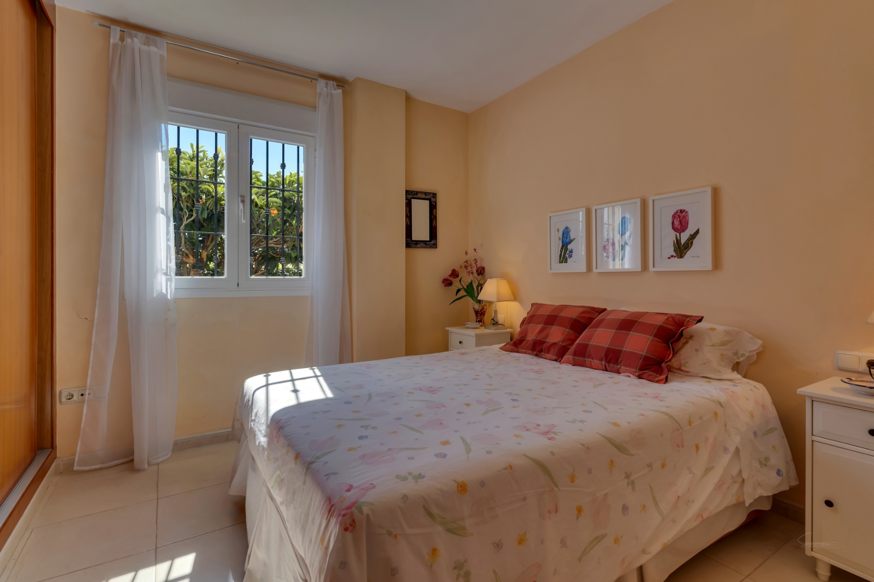Chambre lumineuse aux murs couleur pêche, avec un lit double et une fenêtre à barreaux offrant une vue sur le jardin et une lumière naturelle abondante. Armoire intégrée.