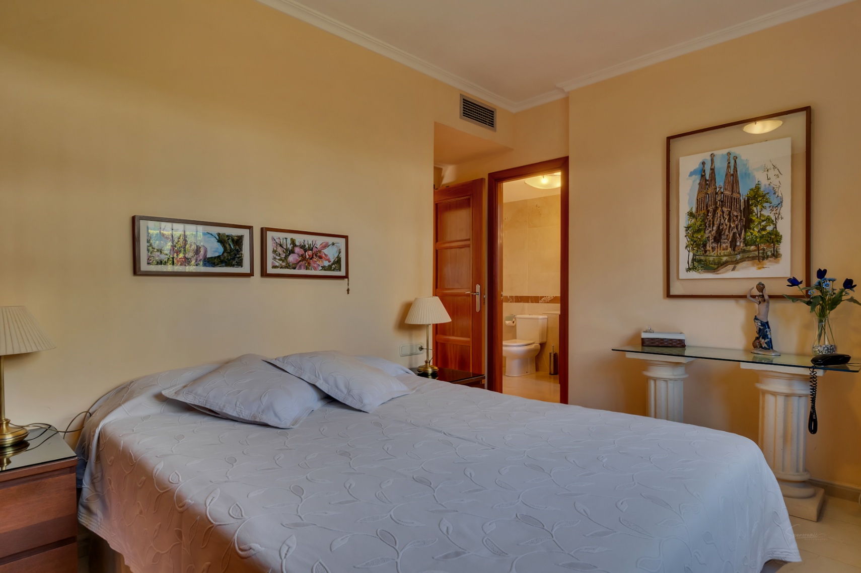 Chambre principale avec lit double, tables de chevet en bois et accès direct à une salle de bain privée. Des œuvres d'art encadrées et une console en verre ornent la pièce.