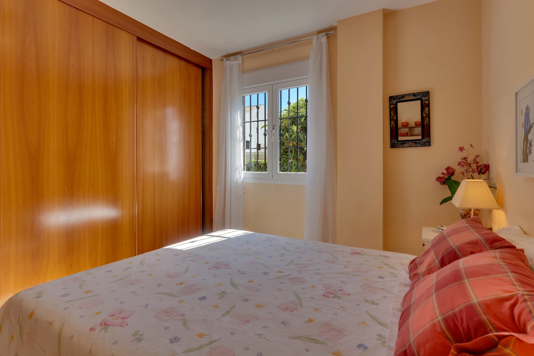 Chambre principale lumineuse avec grande armoire intégrée en bois clair, lit double et fenêtre sécurisée offrant une vue sur la verdure.