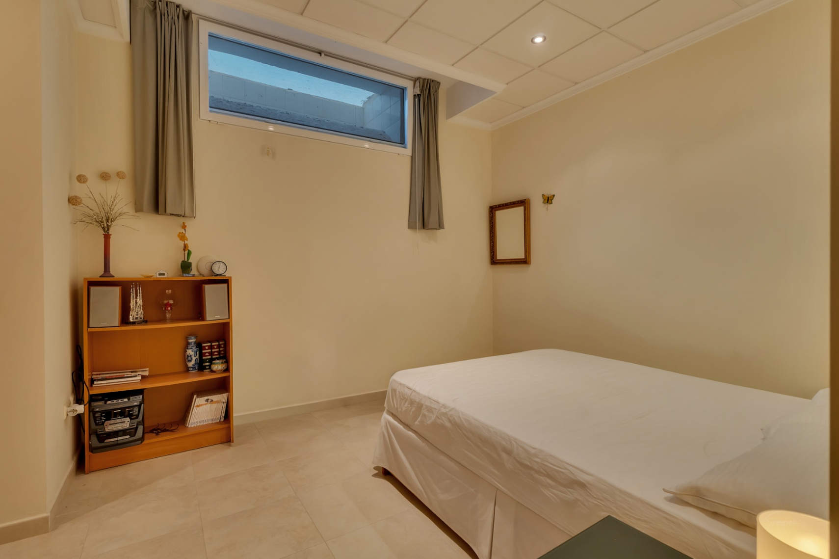Chambre secondaire avec lit simple, carrelage clair et murs crème. Elle dispose d'une étagère en bois, d'un système audio et d'une fenêtre haute avec rideaux gris.