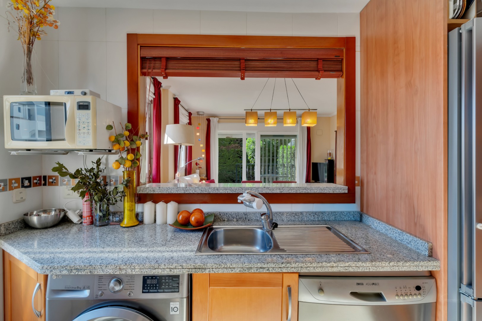 Cuisine fonctionnelle avec plan de travail en granit, évier en acier inoxydable, lave-linge et lave-vaisselle intégrés. Une fenêtre passe-plat en bois s'ouvre sur le salon-salle à manger.