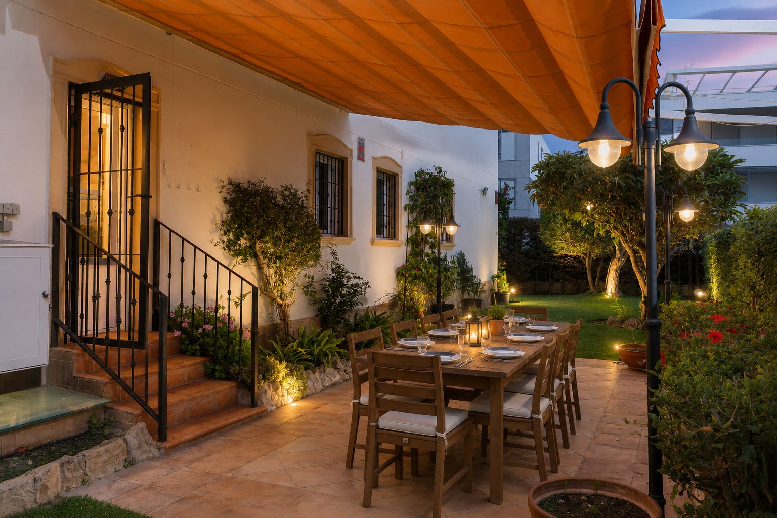 Espace repas extérieur accueillant sur patio carrelé, avec grande table en bois dressée sous un store orange rétractable, entouré d'un jardin illuminé.