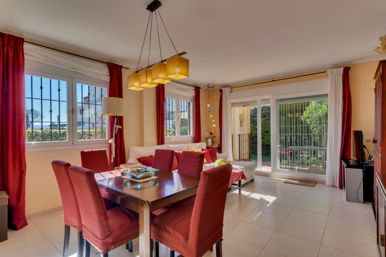 Grand salon-salle à manger avec table en bois pour six, chaises rouges et lumière naturelle abondante. Accès direct au jardin par des portes coulissantes vitrées.