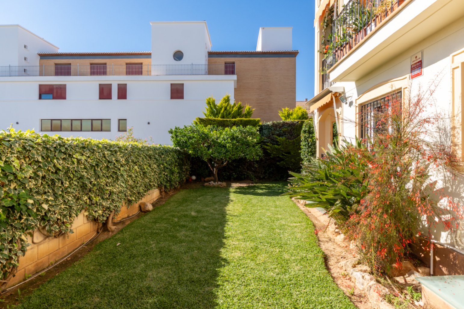 Jardin privé avec pelouse bien entretenue, entouré de haies verdoyantes et d'un petit arbre fruitier. Des bâtiments modernes et une façade avec auvent sont visibles en arrière-plan.
