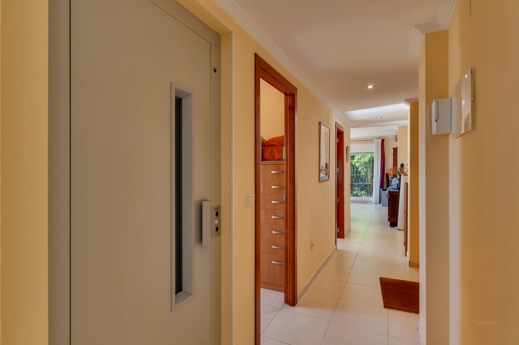 Long couloir aux murs crème et carrelage blanc, doté d'un ascenseur intégré et de plusieurs portes en bois. Au fond, une zone lumineuse avec vue sur le jardin est perceptible.