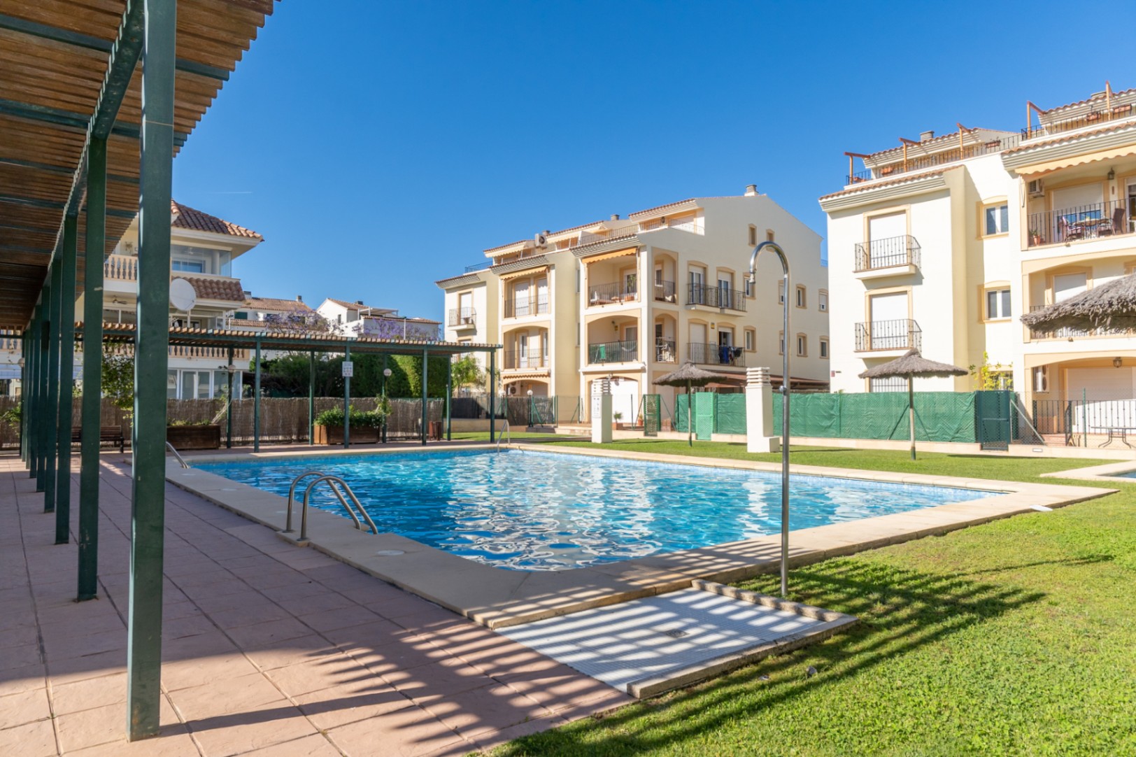 Piscine communautaire d'eau salée avec carrelage bleu, pelouse verte et bâtiments résidentiels. Une pergola en bois et une douche extérieure sont visibles.