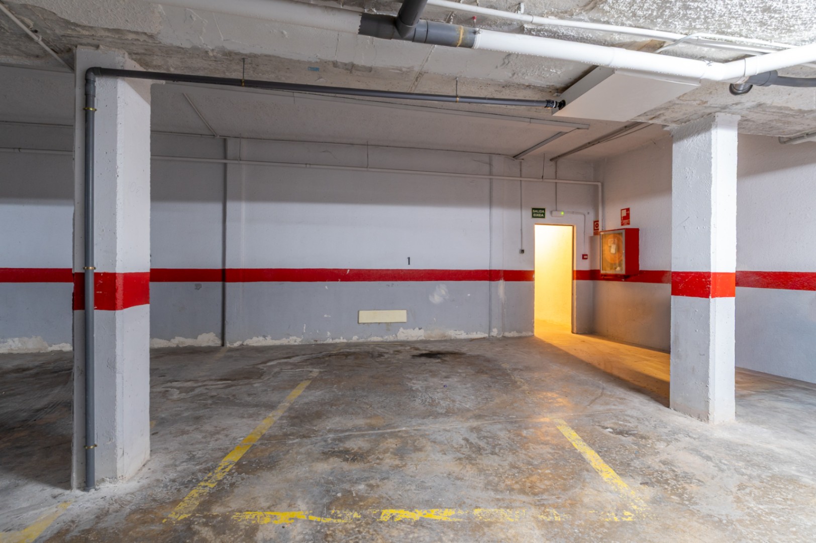 Place de parking souterrain spacieuse avec sol en béton, marquages jaunes et tuyaux apparents. Comprend une porte de sortie éclairée et un équipement anti-incendie.