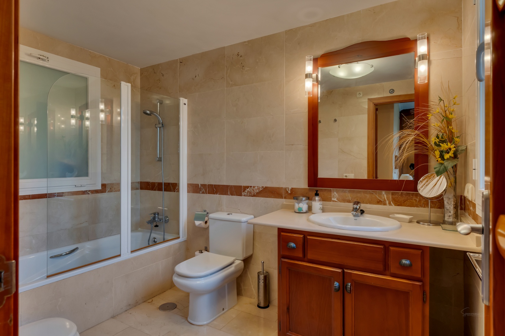 Salle de bain avec carrelage beige clair et bande de marbre, baignoire avec pare-douche vitré, et meuble-lavabo en bois avec miroir éclairé. Une fenêtre dépolie assure l'intimité.