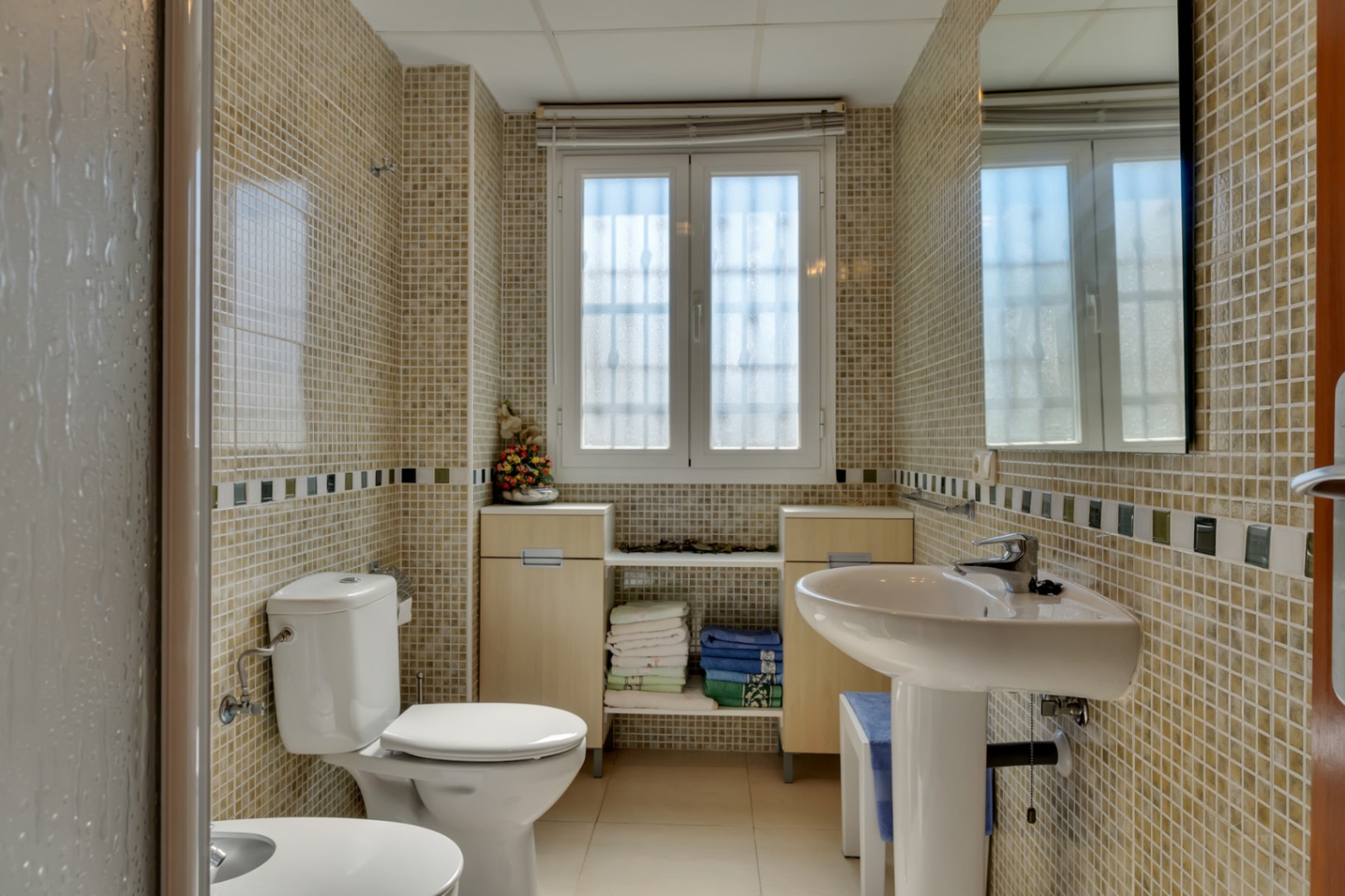 Salle de bain avec murs en mosaïque claire et frise décorative, lavabo sur colonne, toilettes et fenêtre pour la lumière naturelle. Des meubles bas en bois clair offrent du rangement.