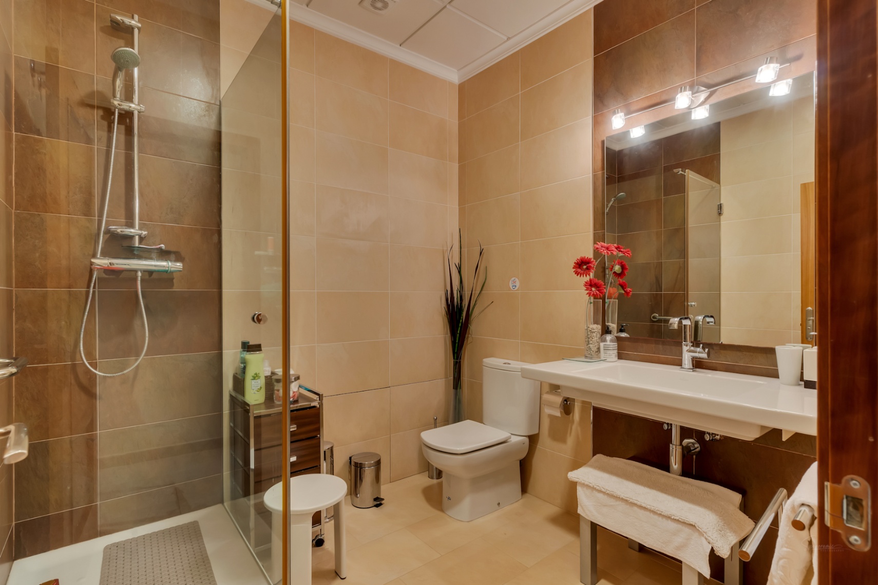 Salle de bain complète avec douche vitrée, carrelage beige et marron, lavabo suspendu et miroir avec éclairage moderne.