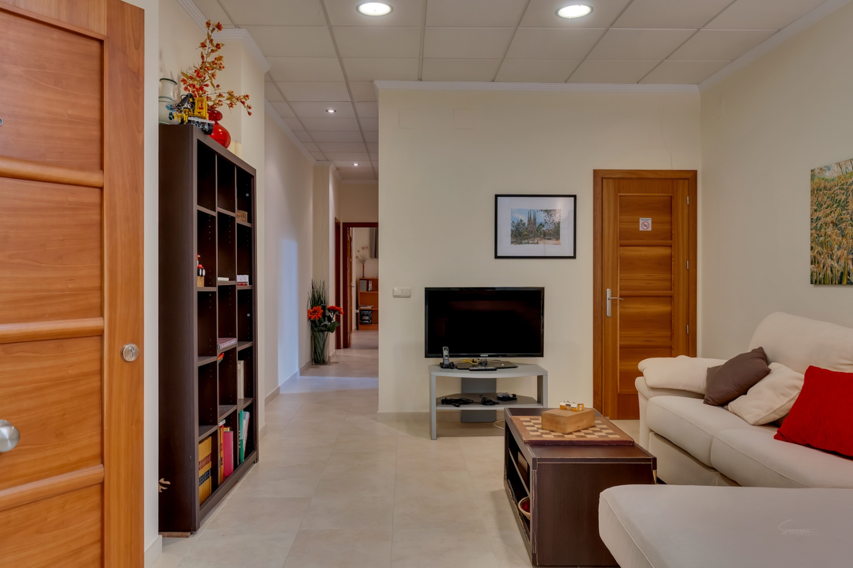 Salon accueillant avec canapé, télévision et table basse avec échiquier. Portes en bois clair, bibliothèque et couloir s'étendant à l'arrière-plan.