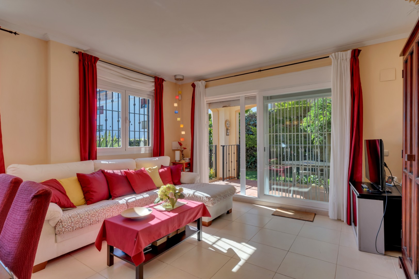 Salon lumineux avec canapé blanc, coussins colorés, sol carrelé et accès direct au jardin privé par des portes coulissantes vitrées.