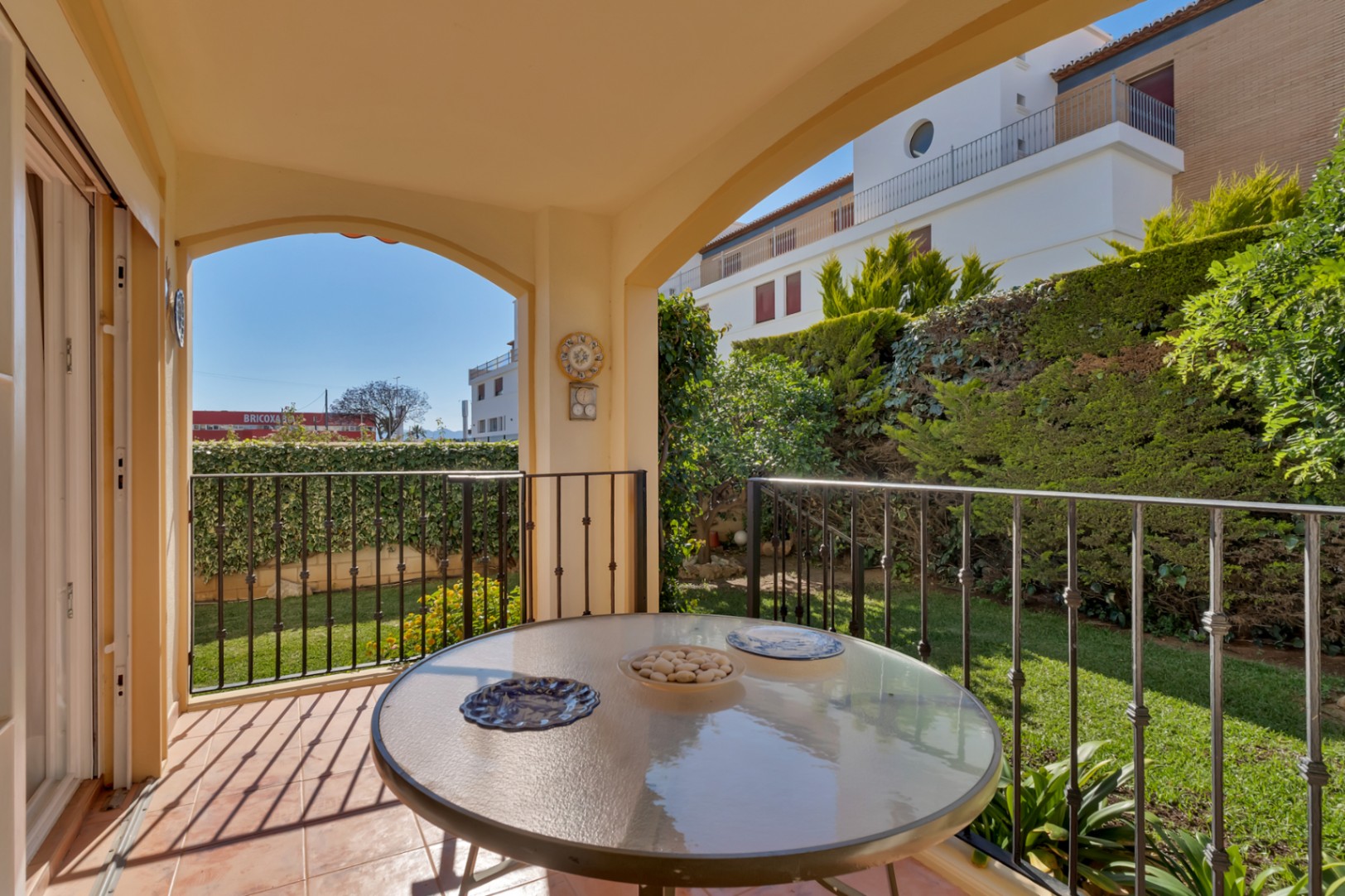 Terrasse couverte avec table en verre et carrelage en terre cuite, offrant une vue directe sur un jardin privé verdoyant avec haies et bâtiments lointains.