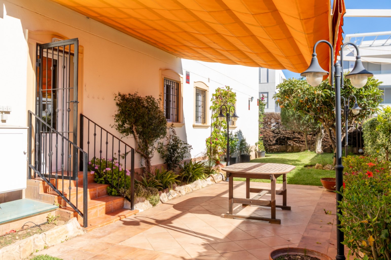 Vaste patio extérieur carrelé de terre cuite, avec une table en bois et un grand store orange rétractable. Accès direct à la maison et au jardin verdoyant.
